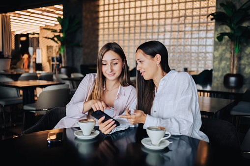 Two gossipy girls are sitting at the table in a coffee shop, having their coffee, looking at social media on the phone while sharing their secrets. Two girls gossiping and looking at the phone.