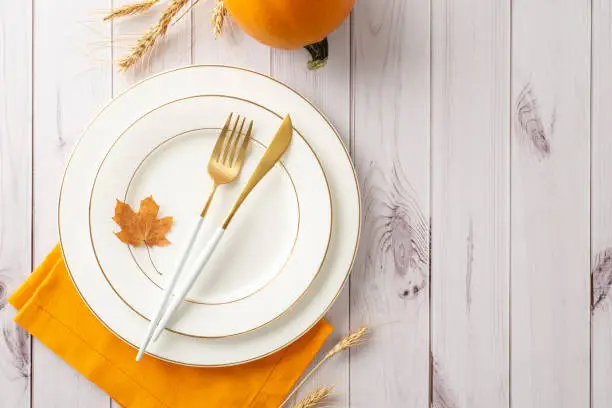 Elevated outlook captures spirit of familial Thanksgiving dinner set. Gilded plates, country-style silverware and fall-themed ornaments on white wooden backdrop, with space for text or advertisements