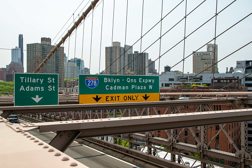 Vibrant road signs on the Brooklyn Bridge with a modern cityscape backdrop, captured through reportage photography in Brooklyn Heights, NY, USA.