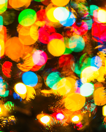 An intimate close-up photograph capturing the high-contrast bokeh of a Christmas tree set against the dark of night. The tree's branches and needles create silhouettes that contrast with the glowing lights in green, yellow, blue, and red. Shot at night with a shallow depth of field during winter, this photo exudes a warm ambiance. It is ideally suited as a texture or wallpaper for festive and holiday-themed settings.