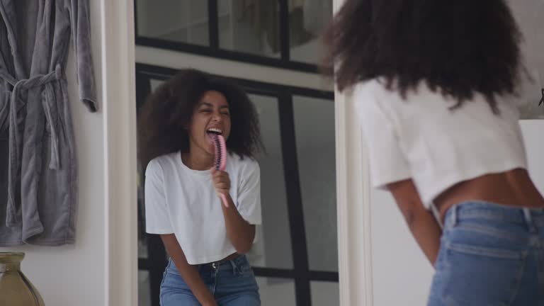 Black Woman Singing in Hairbrush as Microphone. Positive African American female dancing in bathroom looking in the mirror.