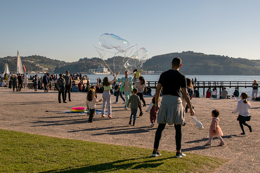 In Lisbon, Portugal, children playfully chase soap bubbles, while joyful people bask in the warmth of a sunny evening at a vibrant park.