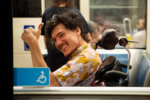 Young asian man using a wheelchair in a subway tram. He has cerebral palsy and wheelchair is motorised. He is looking at camera with a thumb up. Horizontal waist up indoors shot with some copy space. This was taken in Montreal, Quebec, Canada.