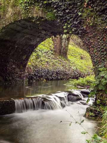 Arch of a medieval bridge where you can see a river with a small waterfall with a silk effect, made with a long exposure.