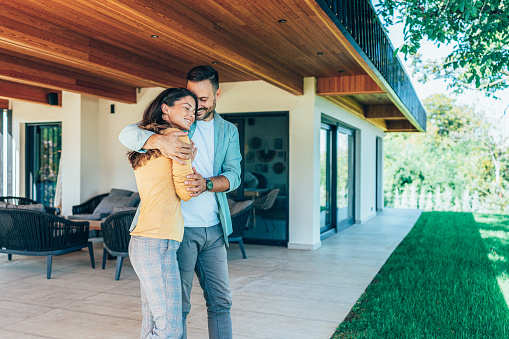 Couple standing in front of their new home. They are both wearing casual clothes and embracing. They are with eyes closed and smiling. The house is contemporary with porchway and a green lawn, wood and glass exterior design