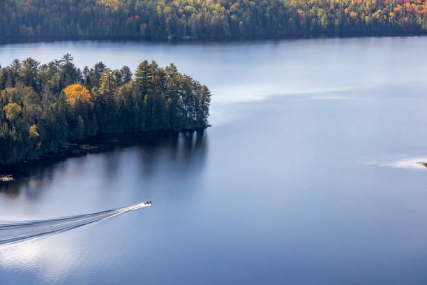 A boat cuts through the reflections of the clouds amidst the Autumn coloured trees, as seen from the top of Centennial Ridges Trail in Algonquin Park, Muskoka, Ontario, Canada.