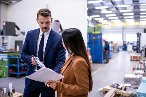 The manager debates with the customer about the actual order of products in their workshop. A white man and an Asian woman are looking at the production. The white man is in a blue business suit with a tie, the visitor is in a coat. Bright manufacturing plant with metalworking machinery. Business meeting, copy space, bright,