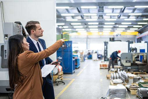 The manager debates with the customer about the actual order of products in their workshop. A white man and an Asian woman are looking at the production. The white man is in a blue business suit with a tie, the visitor is in a coat. Bright manufacturing plant with metalworking machinery. Business meeting, copy space, bright,