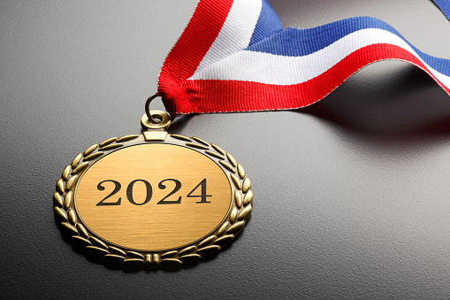 A gold medal engraved with 2024.  A red, white, and blue ribbon is attached to the medal that rests on a background that graduates from gray to black.