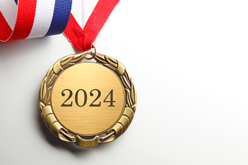 A gold medal engraved with 2024 on it.  A red, white, and blue ribbon is attached to the medal that rests on a white background.