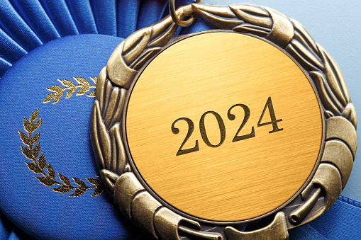 A close up of a gold medal engraved with the year, 2024.  The medal rests next to a blue first place ribbon.