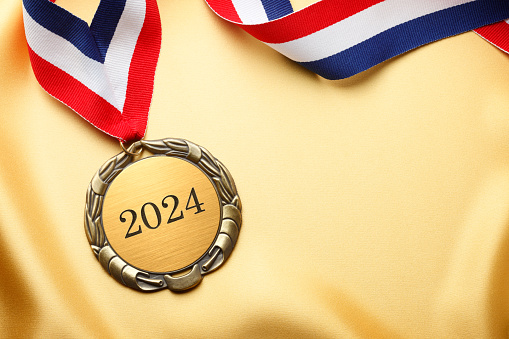 A gold medal engraved with 2024 on it.  A red, white, and blue ribbon is attached to the medal that rests on a piece of gold satin fabric.