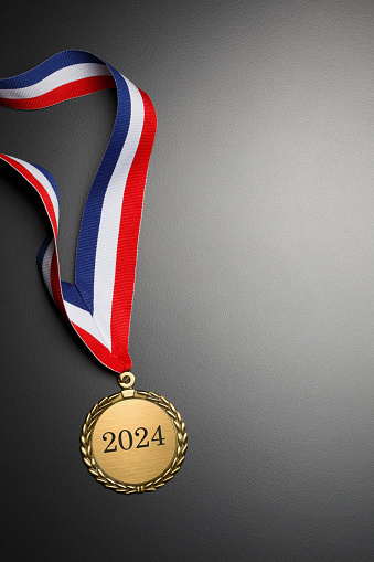 A gold medal engraved with 2024.  A red, white, and blue ribbon is attached to the medal that rests on a background that graduates from gray to black