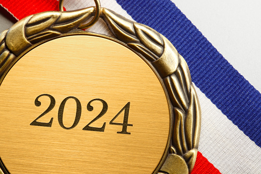 A close up of a gold medal engraved with 2024 on it.  A red, white, and blue ribbon is attached to the medal that rests on a white background.
