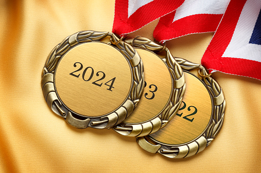 A stack of three gold medals. The top medal is engraved with 2024 and the other two are engraved with 2023 and 2022. A red, white, and blue ribbon is attached to each medal as they rest on a pieces of gold satin fabric.
