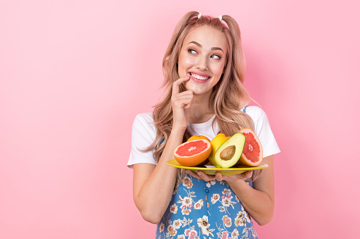 Photo of minded nice woman dressed print overall hold plate with fruits look empty space finger teeth isolated on pink color background.