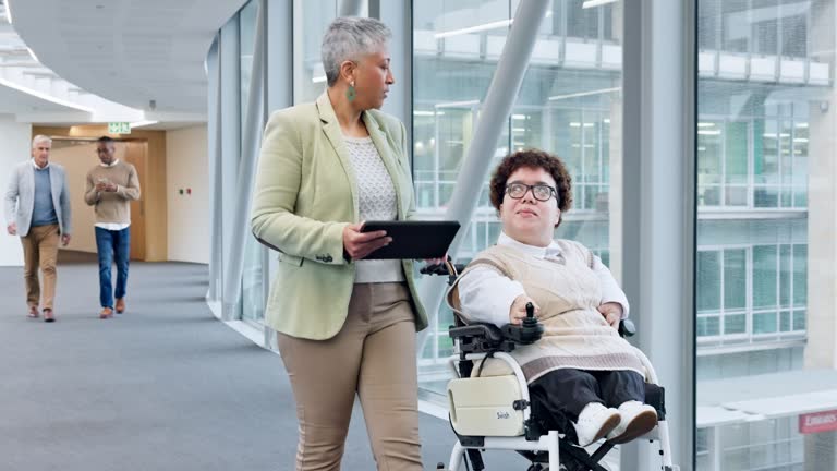 Business women, wheelchair and discussion in office hallway on tablet, schedule or teamwork in workplace. Mature manager, partnership and person with disability for ideas, talk or digital touchscreen