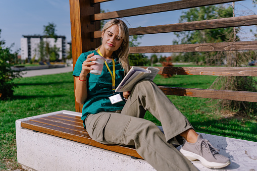 Young Caucasian female nurse in scrubs, peacefully enjoying her book and coffee on a sunny day.