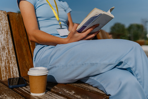 Close-up of a young Caucasian woman`s hand holding a book, while sitting on a park bench. She has a coffee sub next to her and is wearing scrubs.