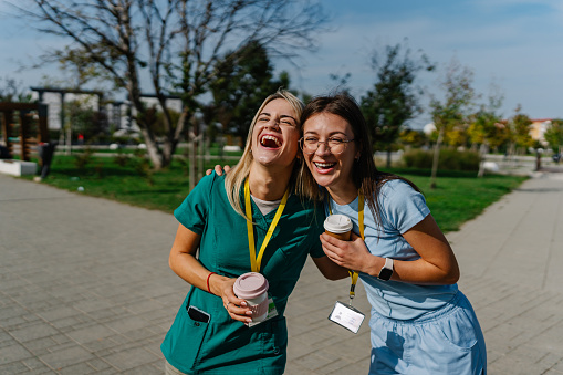 Two young Caucasian female nurses in scrubs cheerfully embracing on a sunny day. They are enjoying their coffee and laughing.