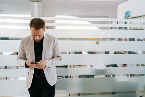Medium shot portrait of busy businessman in suit using mobile phone standing in light hallway corridor of modern business center. Business man in suit holding smartphone, reading sms from colleagues