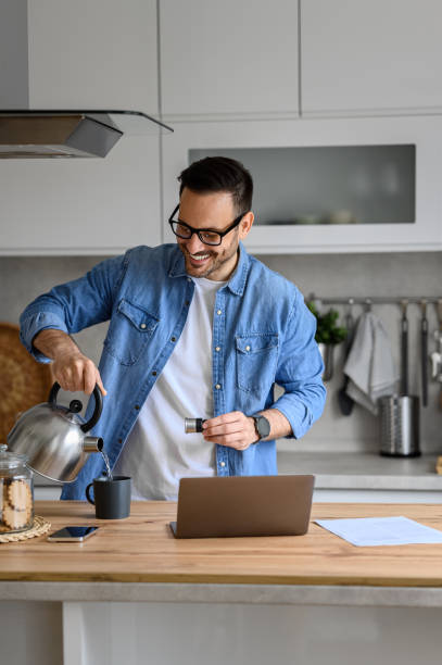 istock Smiling businessman pouring hot water in coffee cup while working over laptop on kitchen counter at home 1720235896