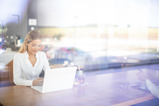 Young confident businesswoman sitting in cafeteria in office building and working on laptop. She is wearing business casual clothes and there is glass of hot tea on the table. Photo taken through the window