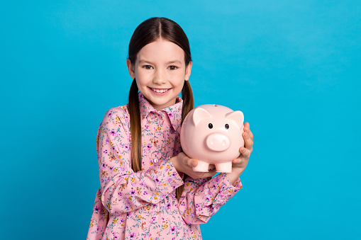 Photo of cute young funny schoolgirl kid brunette hold piggy bank collect money cash savings for dream isolated on blue color background.