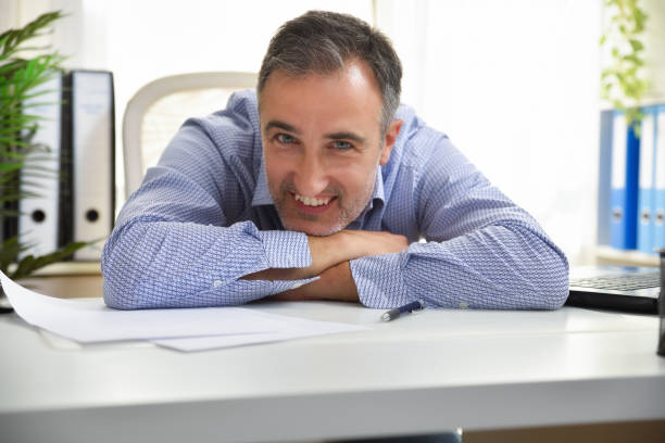 Freelance in blue shirt smiling and leaning on an white office table with papers and folders in the background