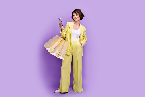 Full length photo of pretty shiny woman dressed yellow jacket holding shoppers texting apple samsung iphone isolated purple color background