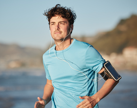 Running, music streaming and man headphones run by the beach and ocean with audio. Mobile radio, web podcast or training song of a runner athlete listening to a track on an outdoor run by the sea