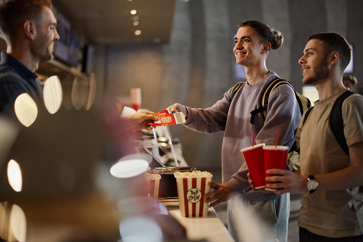 Happy male friends buying movie tickets, snack and drinks at concession stand in cinema.