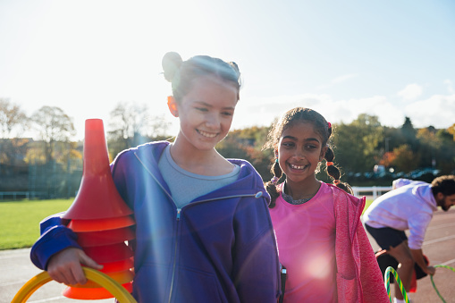 A close up shot of a two female children standing together on an athletics track outside Wentworth Leisure Centre in Hexham, North East England. They are wearing sports clothing and gathering sports equipment before they begin to run. It is a sunny day and in the background is a sports field and trees beyond. The coach is also in the background.

Video also available of this scenario.