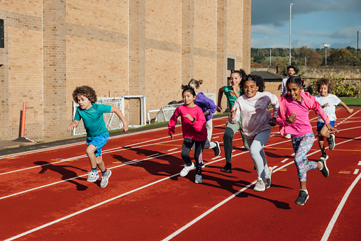 A wide shot of a group of children and a coach running along an athletics track outside Wentworth Leisure Centre in Hexham, North East England. They are wearing sports clothing and some of them are smiling and laughing ad they run. It is a sunny day.

Video also available of this scenario.