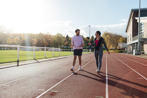 A wide shot of two coaches walking side by side along an athletics track outside Wentworth Leisure Centre in Hexham, North East England. They are wearing sports clothing and discussing. It is a sunny day.

Video also available of this scenario.