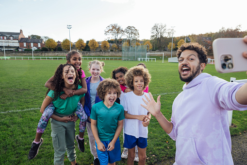 A group of children and a mid adult man coach standing together by an athletics track outside Wentworth Leisure Centre in Hexham, North East England. They are wearing sports clothing and taking a selfie on a smartphone before they begin to run. It is a sunny day and in the background is a sports field and trees beyond.

Video also available of this scenario.