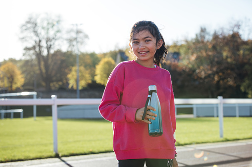 Front view three quarter length portrait of a young female child standing on an athletics track outside Wentworth Leisure Centre in Hexham, North East England. She is wearing sports clothing, holding a reusable water bottle and smiling while looking at the camera. It is a sunny day and in the background is a sports field.

Video also available of this scenario.