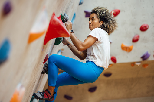 A medium close up side view of a determined woman who is challenging herself and taking on the biggest wall in the indoor climbing centre in an indoor climbing centre in Newcastle upon Tyne. She looks deep in concentration and she also has her hands chalked for grip and is wearing specialty shoes for climbing.