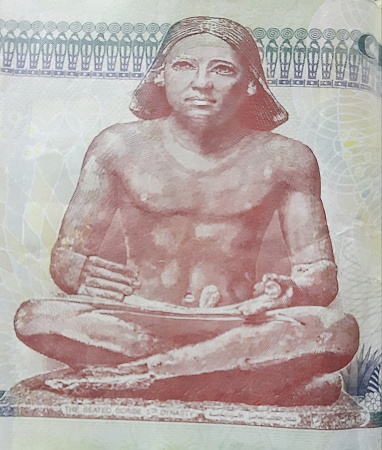 Egypt, Kafr El-Sheikh, a brown statue of the ancient Egyptian writer sitting cross-legged  (ancient egyptian sculpture of seated scribe) on a 200-pound Egyptian banknote, with ancient Pharaonic floral decorations behind him.