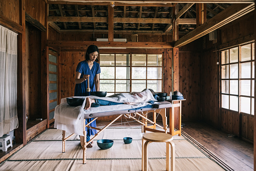 Japanese alternative therapist concentrates on the gentle vibrations emanating from the sound bowls as they work their therapeutic magic on her client.