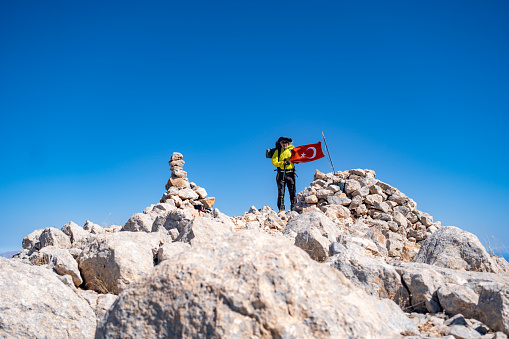 Elderly man with backpack hiking on top of mountain.Hiking pole in hand.Man with backpack.Full equipment.Mediterranean area,mountain area.Clear blue sky.Space for text.Flag at the top. Extreme terrain. Extreme sports. High vitality, active lifestyle. High spirit of adventure.