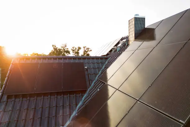 Dark solar panels on the rooftop of a single-family home. The sun is setting over the roof creating a lens flare.