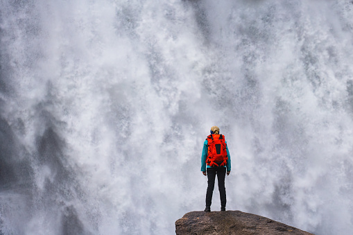 rear view of one woman with huge backpack standing alone on rock close to big powerful waterfall