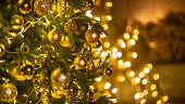 Golden baubles with fairy lights on Christmas tree at home. Holiday ornament with balls and lamp garland on fir tree. Festive decoration with sparkling details and lights on blurry background
