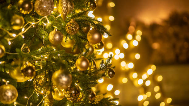 golden baubles with fairy lights on christmas tree at home. holiday ornament with balls and lamp garland on fir tree. festive decoration with sparkling details and lights on blurry background - on branch imagens e fotografias de stock