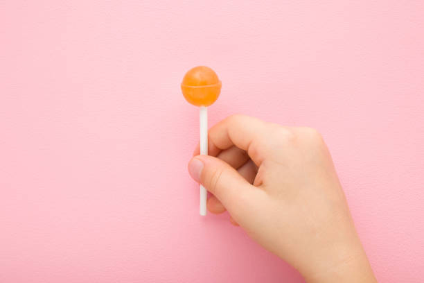 dedos de criança pequena segurando pirulito laranja no pau no fundo rosa claro da mesa. cor pastel. doces doces. closeup. vista de cima para baixo. - hard candy fotos - fotografias e filmes do acervo