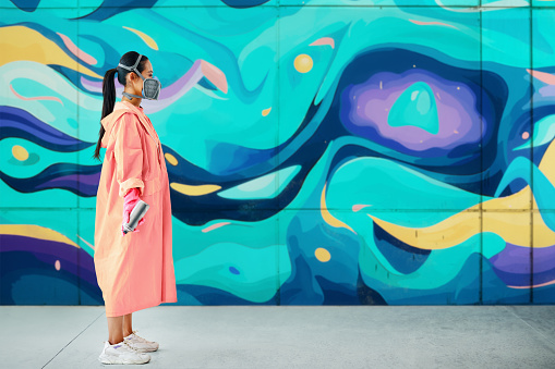 Profile view of female street artist in respirator mask on background of the wall with her colorful paintings with copy space. Urban art concept