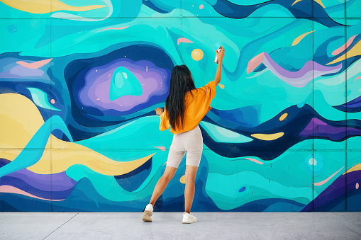 Female street artist painting bright colorful graffiti on wall. Modern art, urban concept. Full length portrait