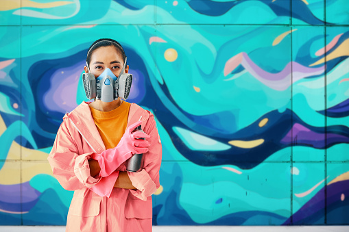 Graffiti female artist painter in respirator mask standing near the wall with her paintings looking to camera with copy space. Street art concept