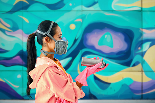 Profile view of female street artist in respirator mask on background of the wall with her colorful paintings with copy space. Urban art concept